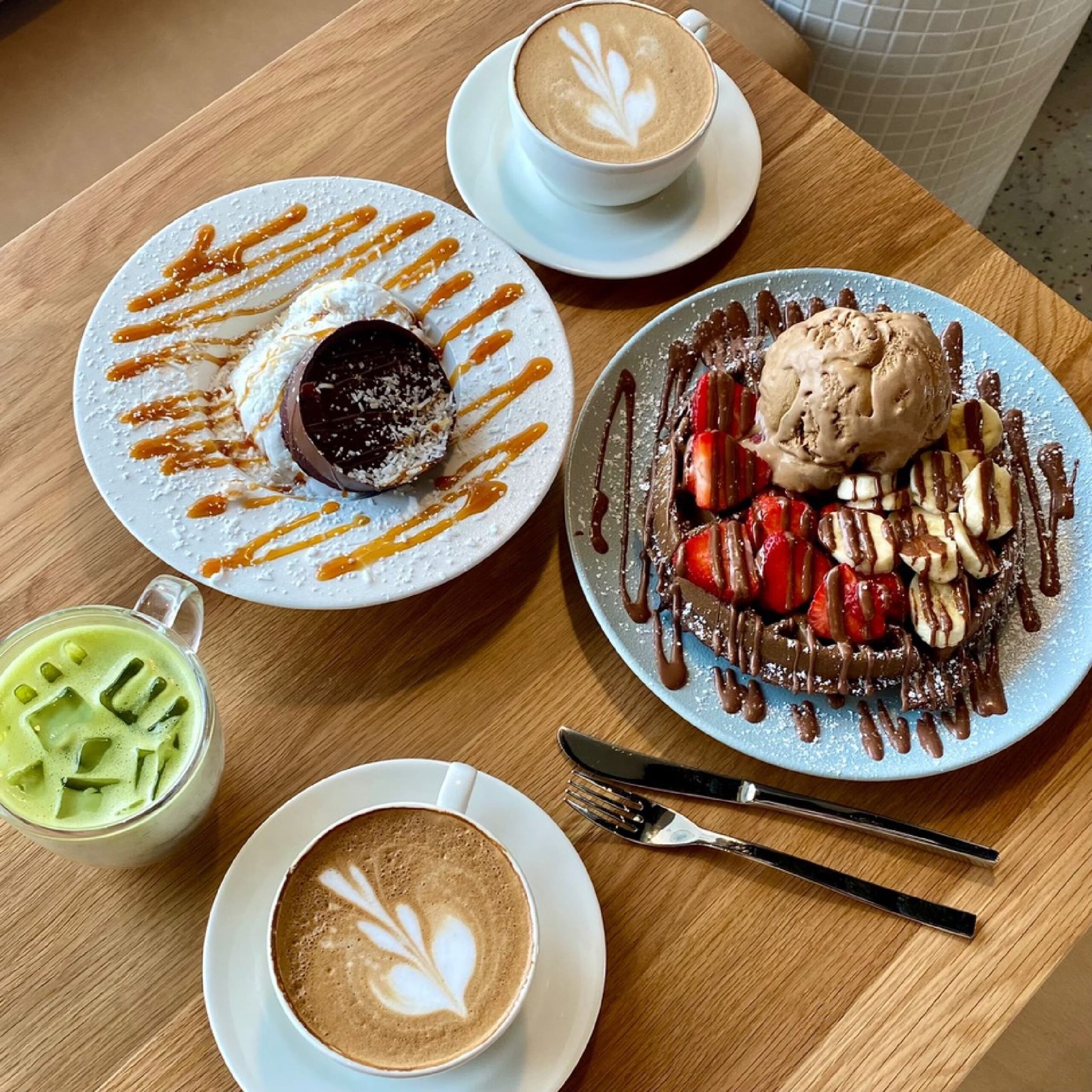Two cappuccinos, one match latte, and two full plate desserts on a table