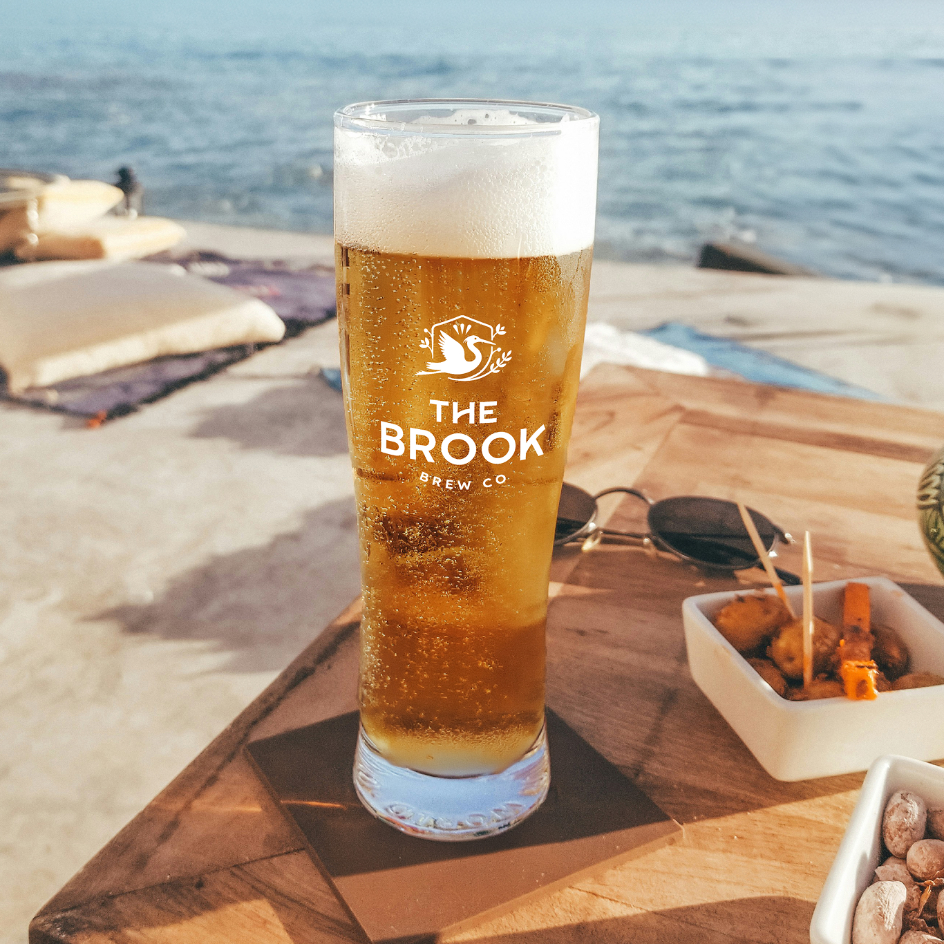 A glass of beer with "The Brook Brew Co" logo on a wooden table by the sea, next to nibbles and sunglasses.