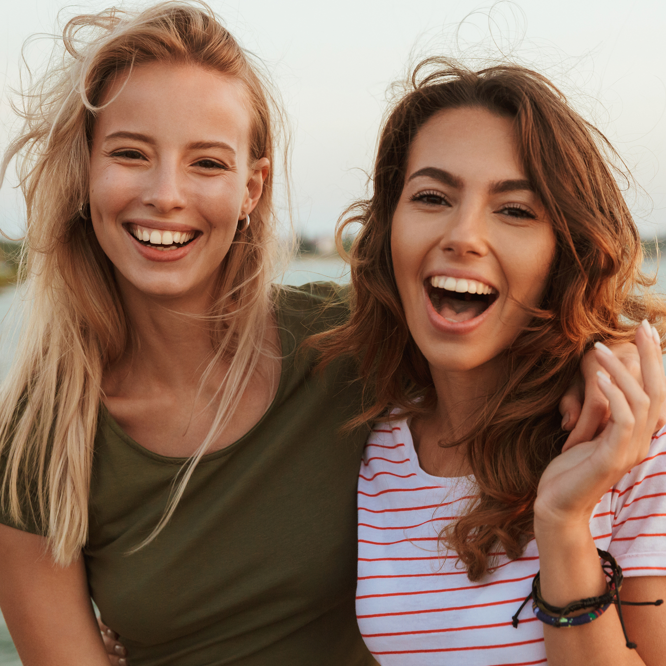 Two young women smiling outdoors, one in a green shirt and the other in a striped top, standing close together.