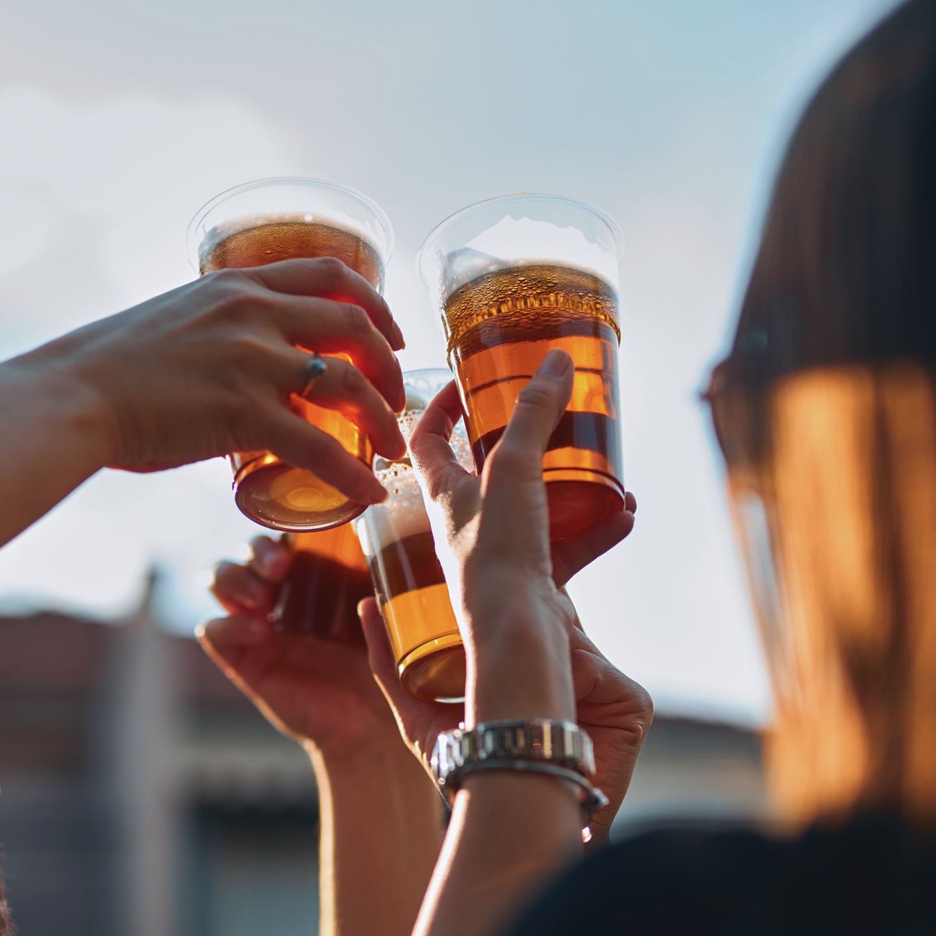 Four people hold up plastic cups filled with beer, making a toast outdoors in natural daylight.