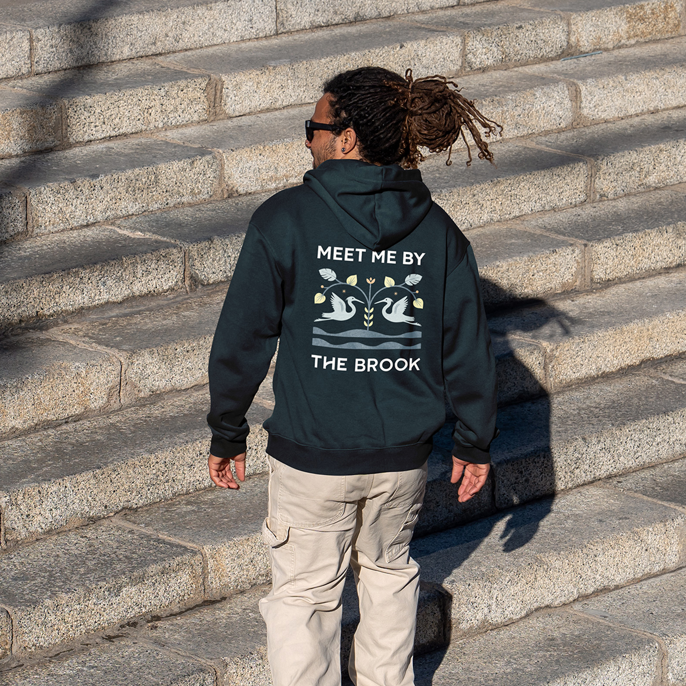 A person wearing a black hoodie with "MEET ME BY THE BROOK" printed on the back, walking up stone steps.