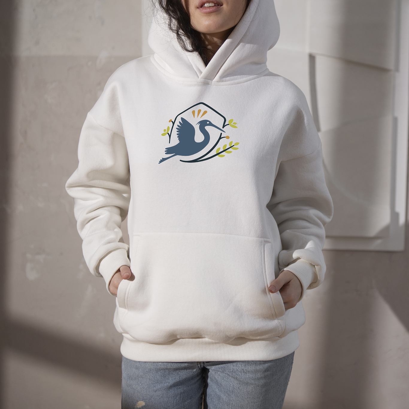 Person wearing a white hoodie with a graphic of a bird holding a branch, standing indoors against a neutral background.