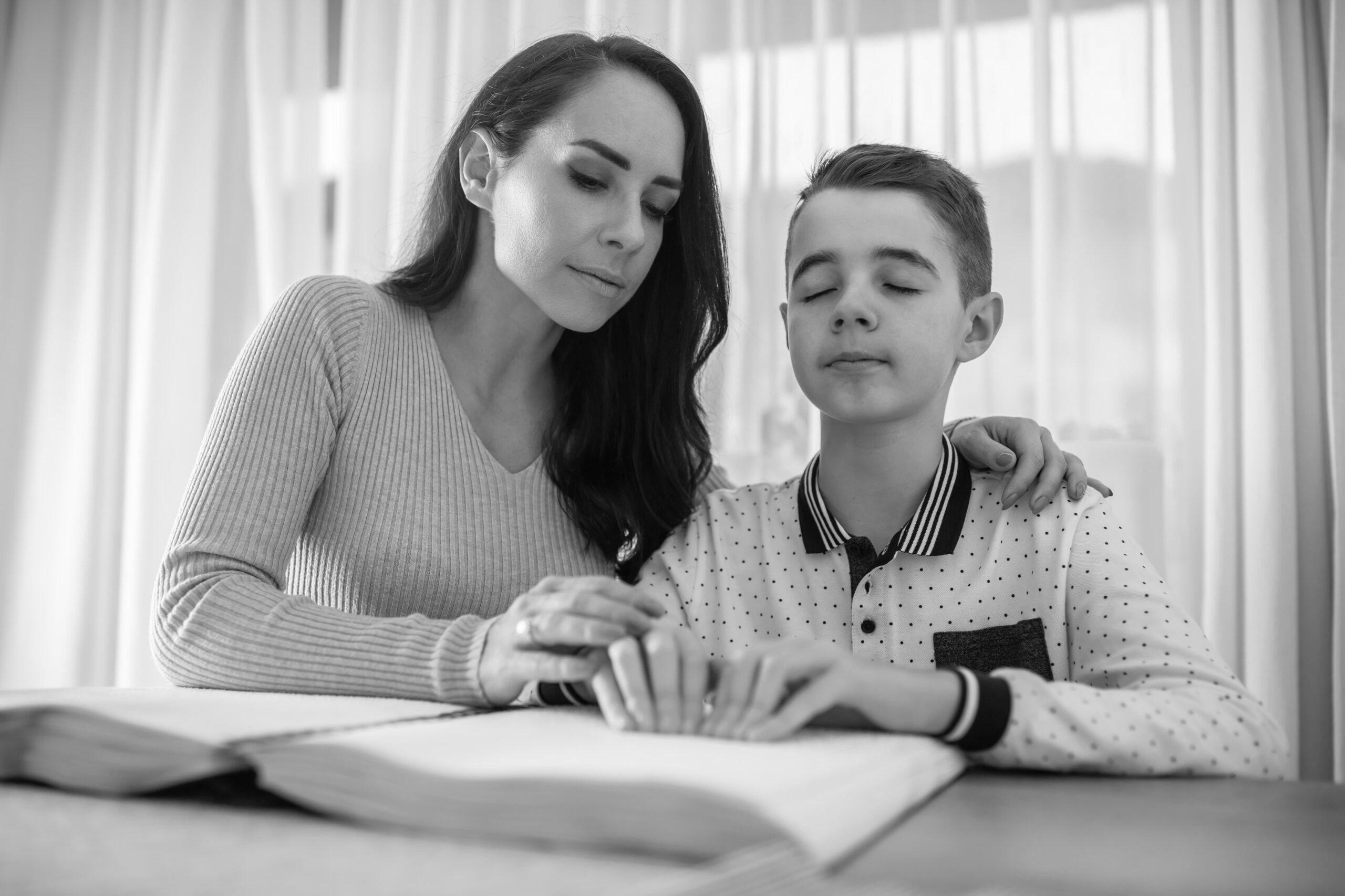 A woman assists a boy in reading Braille from a book at a table together by a window.
