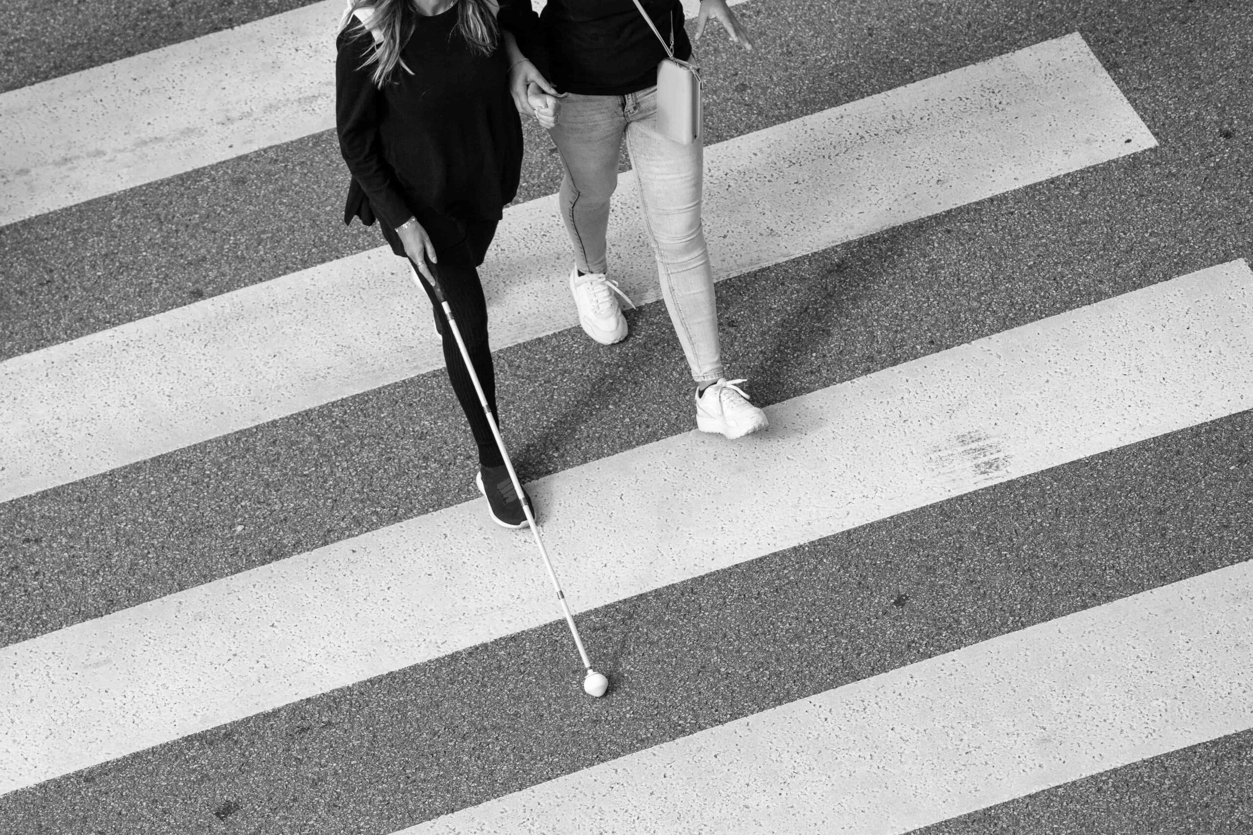 Two people cross a road; one uses a white cane for navigation and the other walks closely beside them.