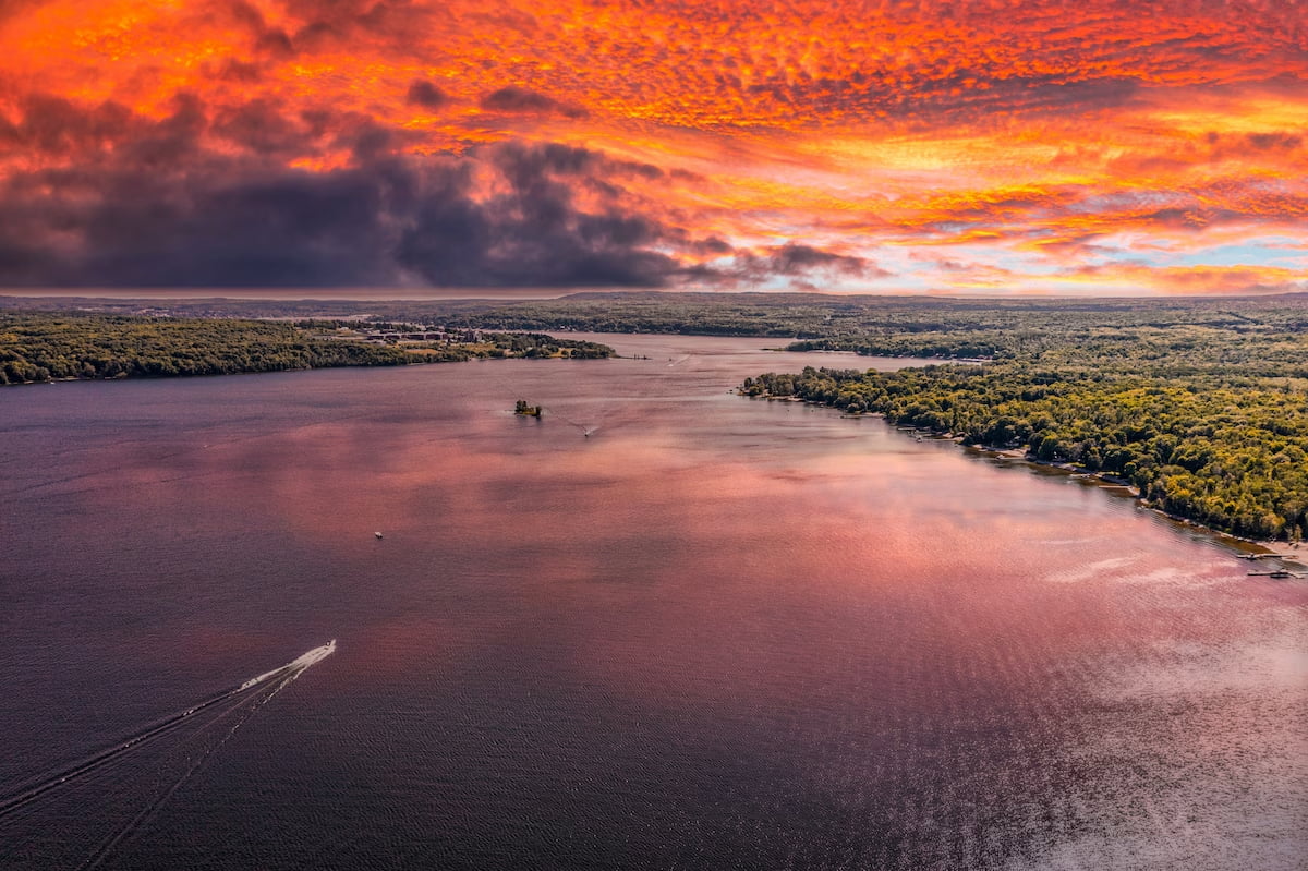 A boat moves across a wide lake under a vivid orange and red sunset sky with scattered clouds and forested shores.