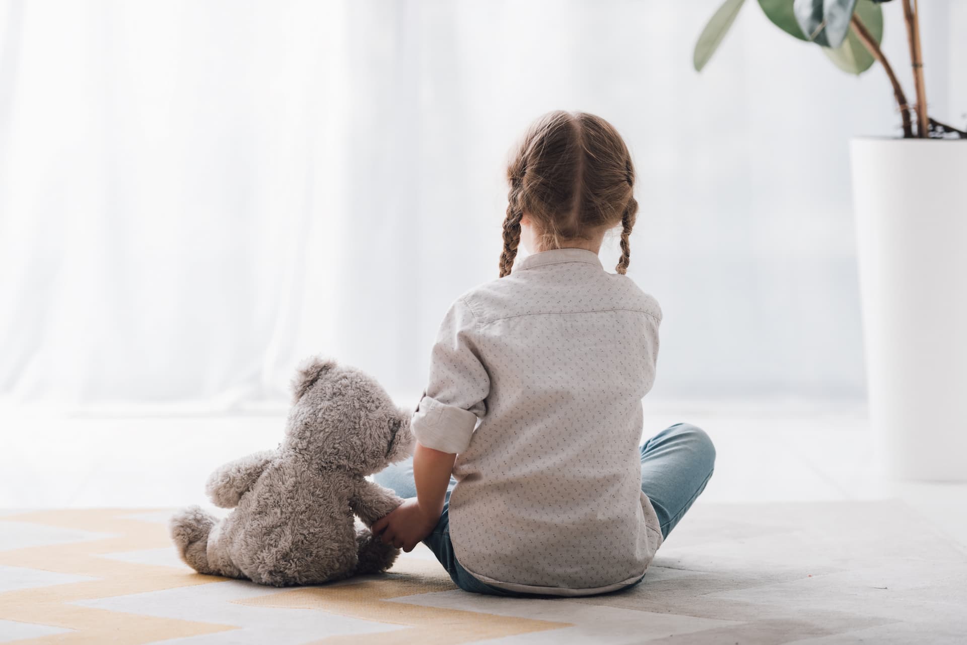 Young child with plaited hair sits on a carpet next to a stuffed teddy bear, facing away from the camera.