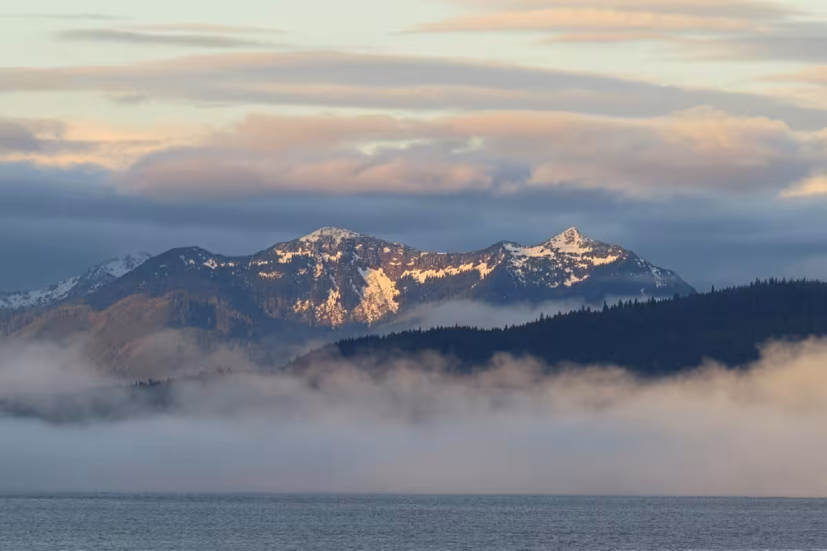 Snow-capped mountains rise above a misty lake with clouds and forested hills in the background.