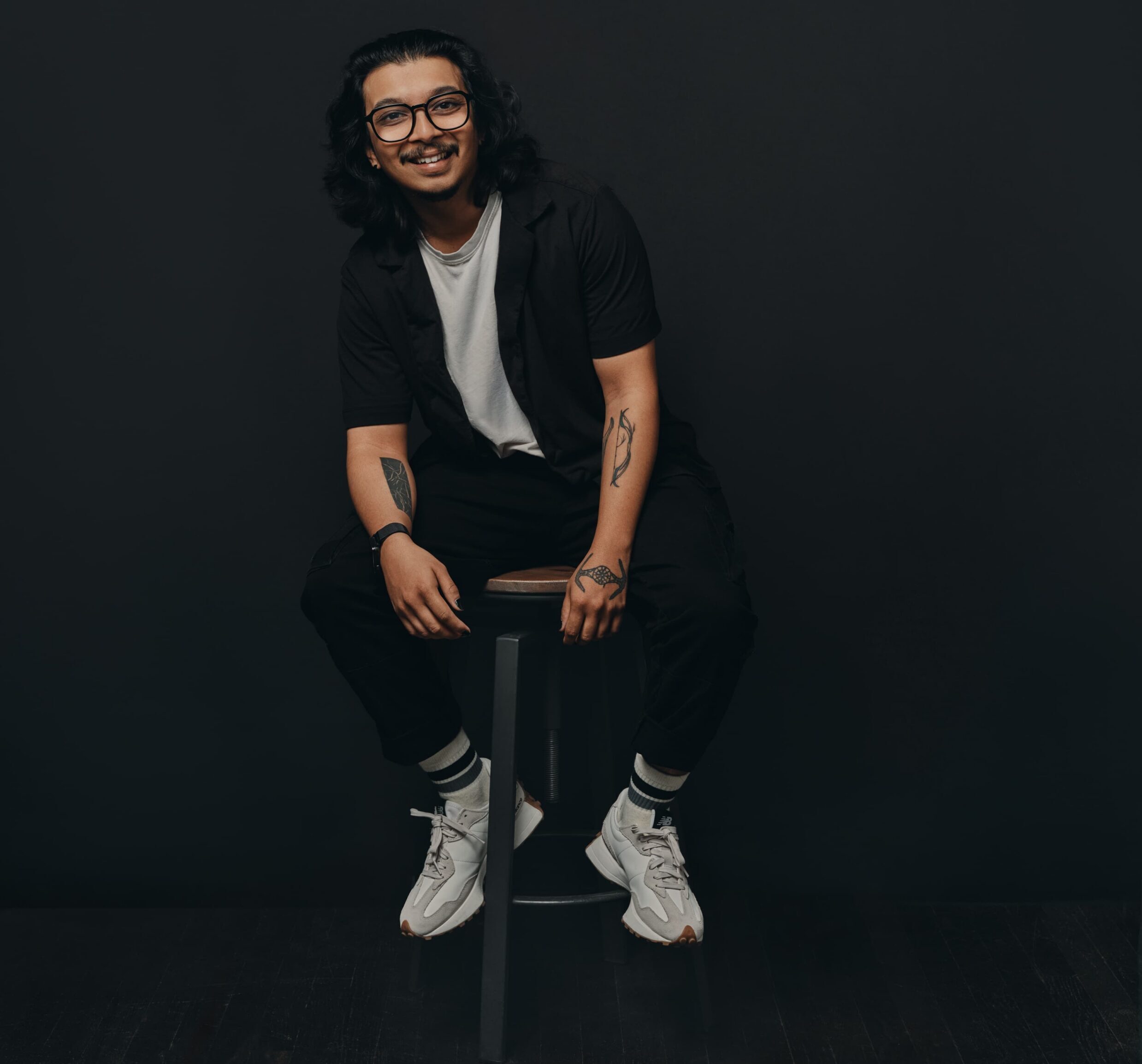 Person with long hair and glasses sits on a stool against a dark background, smiling, hands resting on knees.