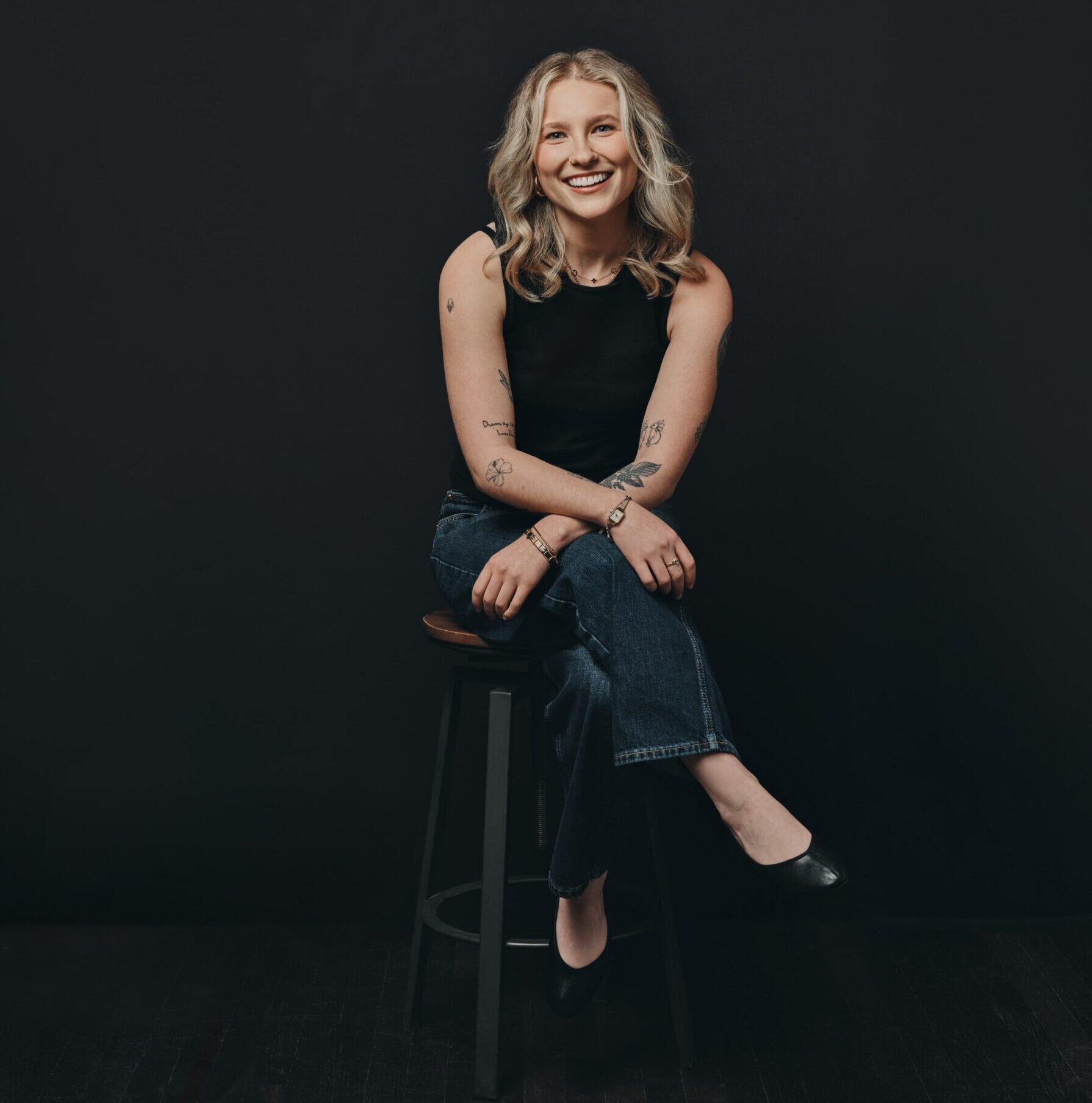 A woman with blonde hair, wearing a black sleeveless top and jeans, sits on a stool against a dark background, smiling.