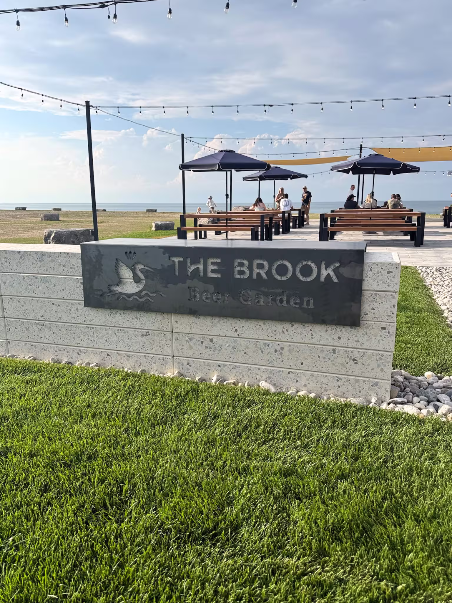 Outdoor seating area with tables and parasols near a sign reading “The Brook Beer Garden” on a sunny day.