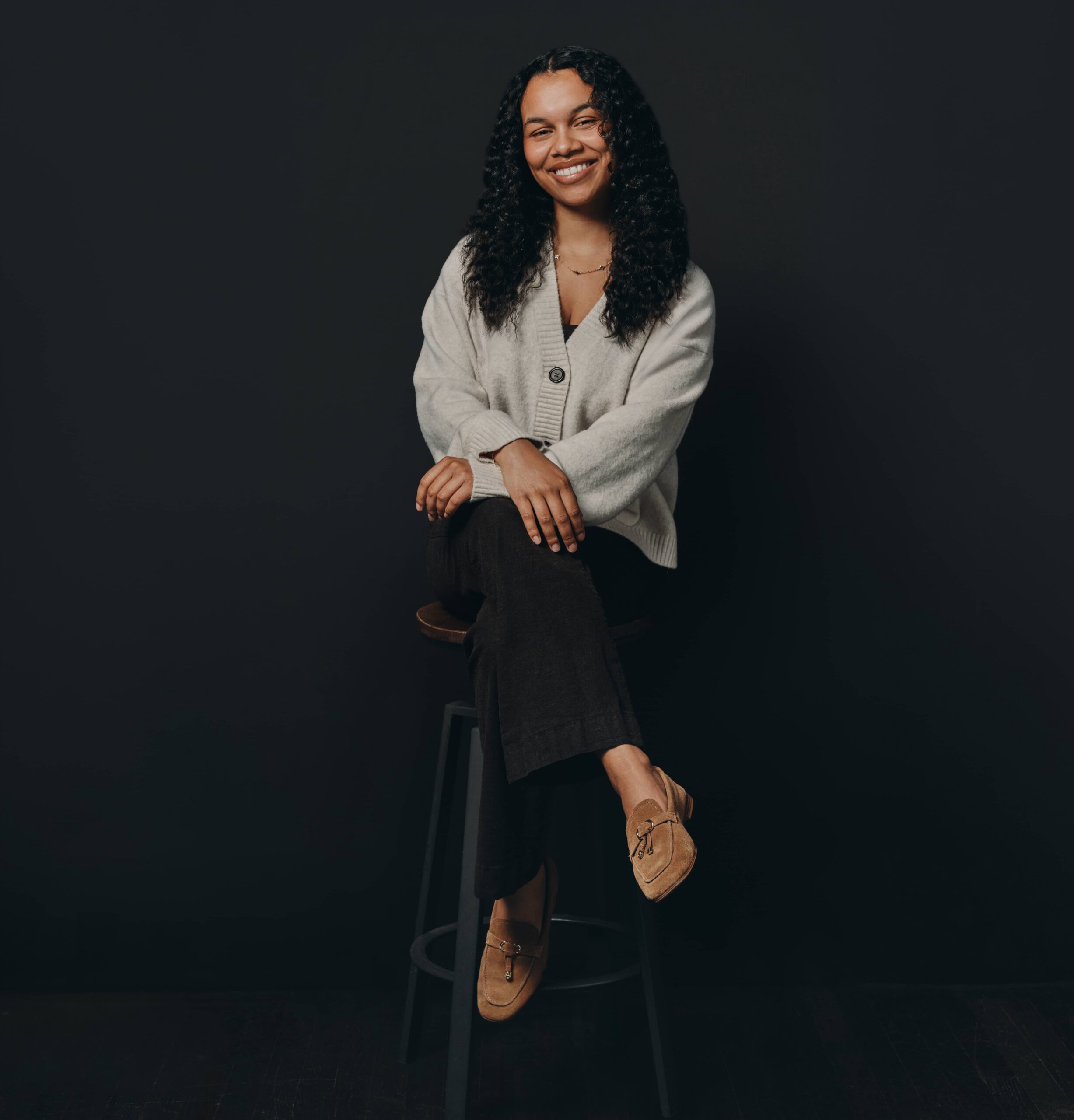Woman with curly hair sits on a stool, wearing a grey jumper, black trousers, and brown shoes against a dark background.