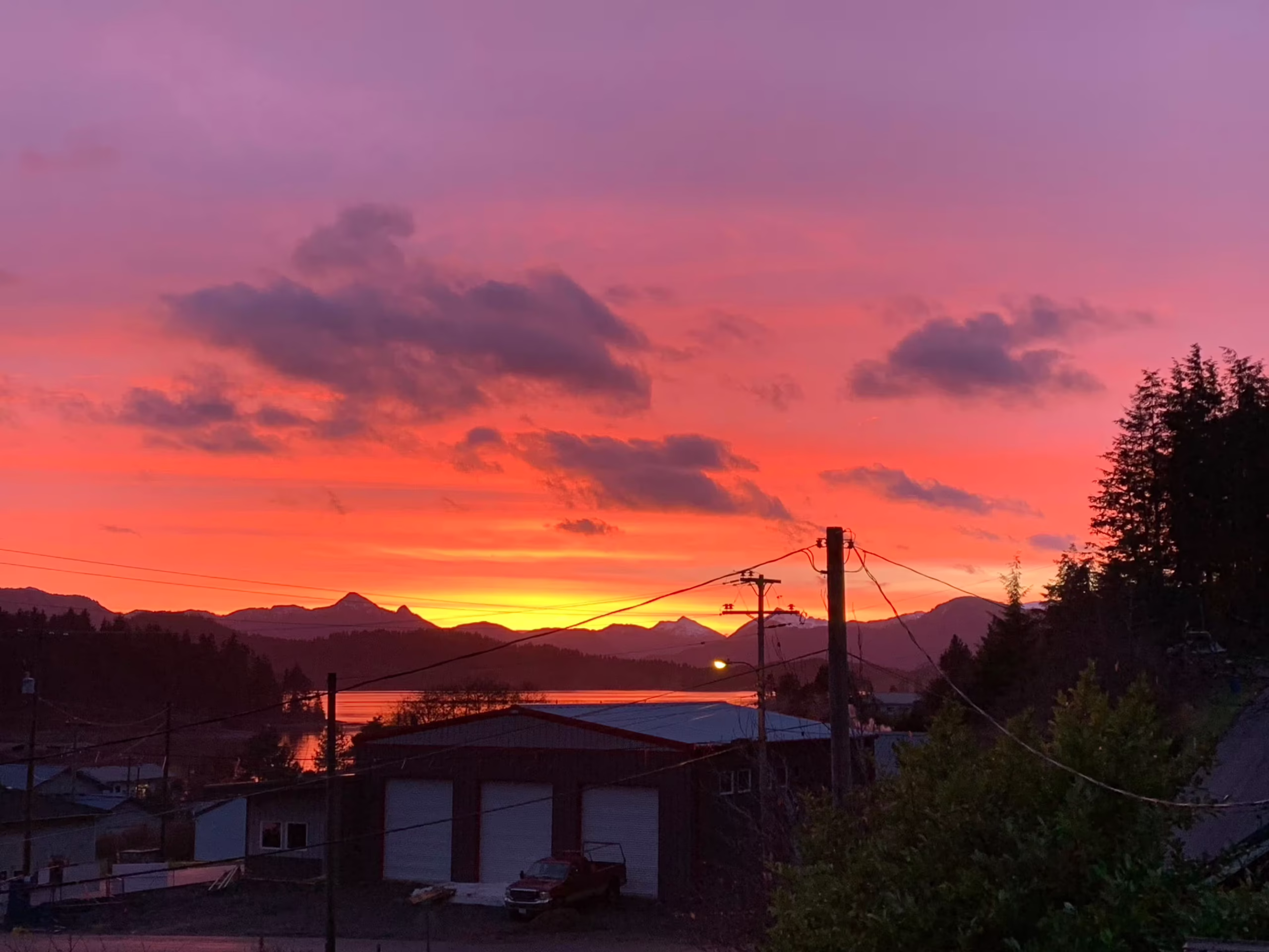 Vivid orange and pink sunset over mountains, water, and a small town with buildings and trees in the foreground.