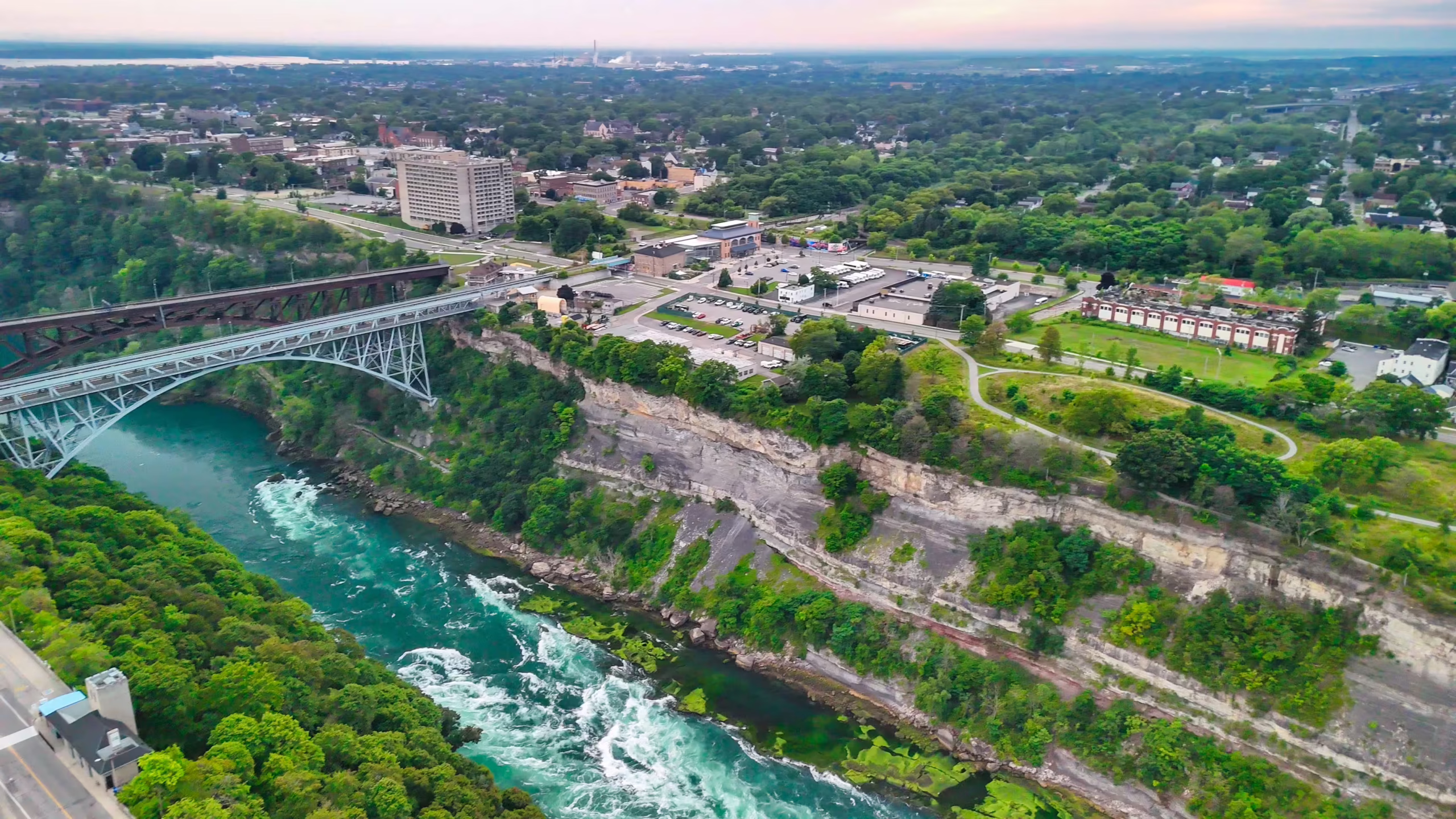 Aerial view of a bridge spanning a river gorge, with buildings, trees, and a cityscape in the background.