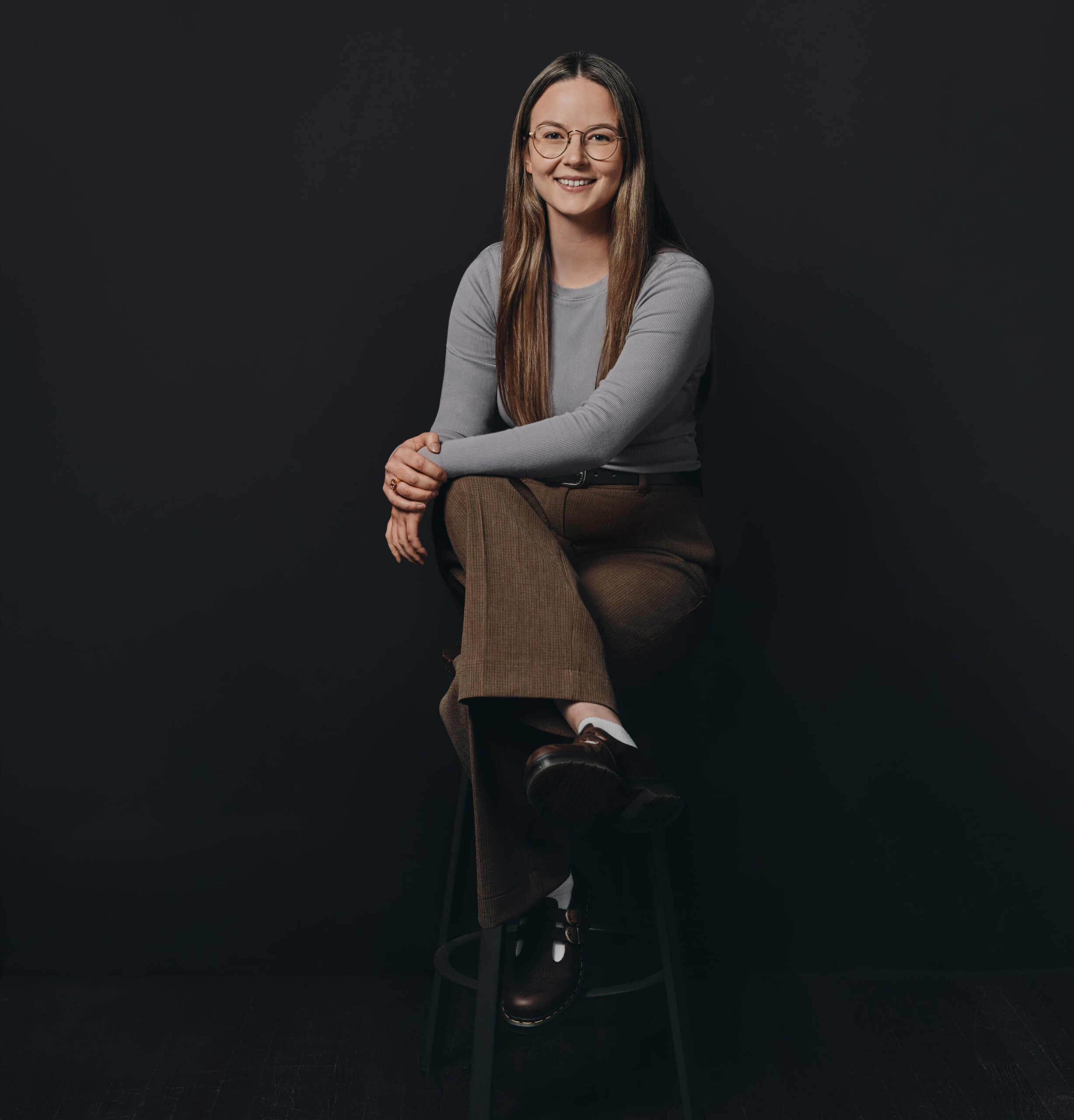 A woman with long hair and glasses sits on a stool against a dark background, smiling at the camera.