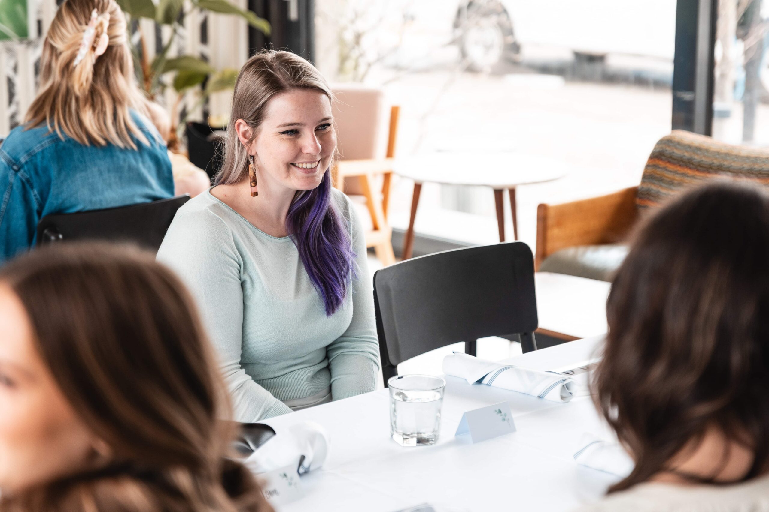 Woman with purple hair sits at a table set for a meal, smiling, with other people and empty chairs around her.