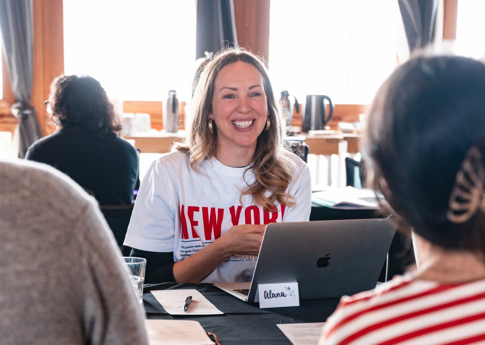 Woman smiling at a table with her laptop, repping “New York,” in a casual marketing agency meeting setting.