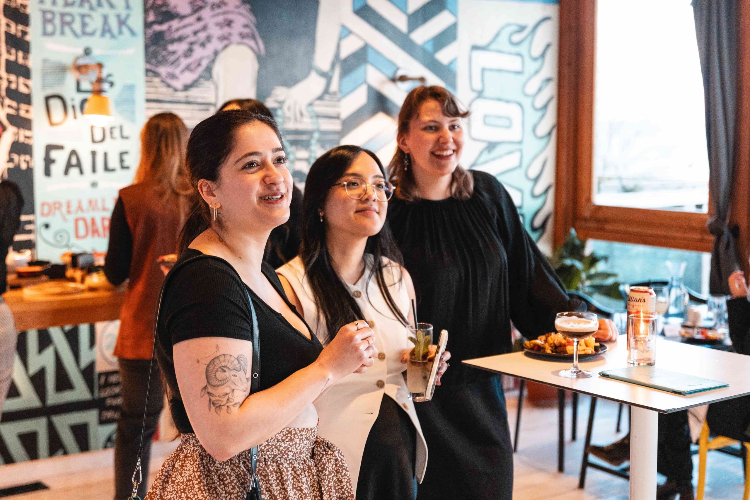 Three women from a marketing agency stand together at a social event, smiling with art-covered walls in the background.