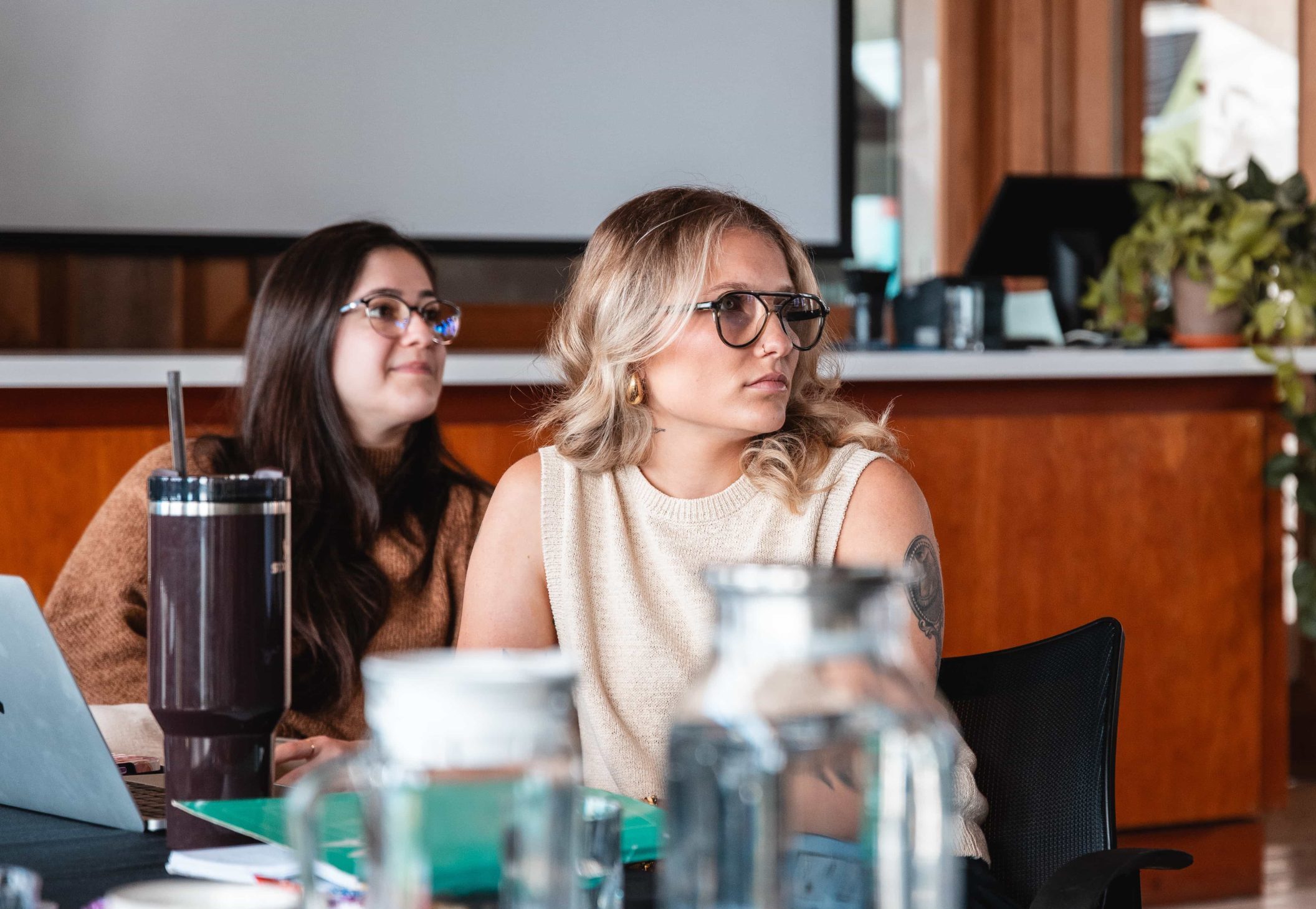 Two women with glasses sit at a table in a modern office, listening attentively. Contact us for more info.