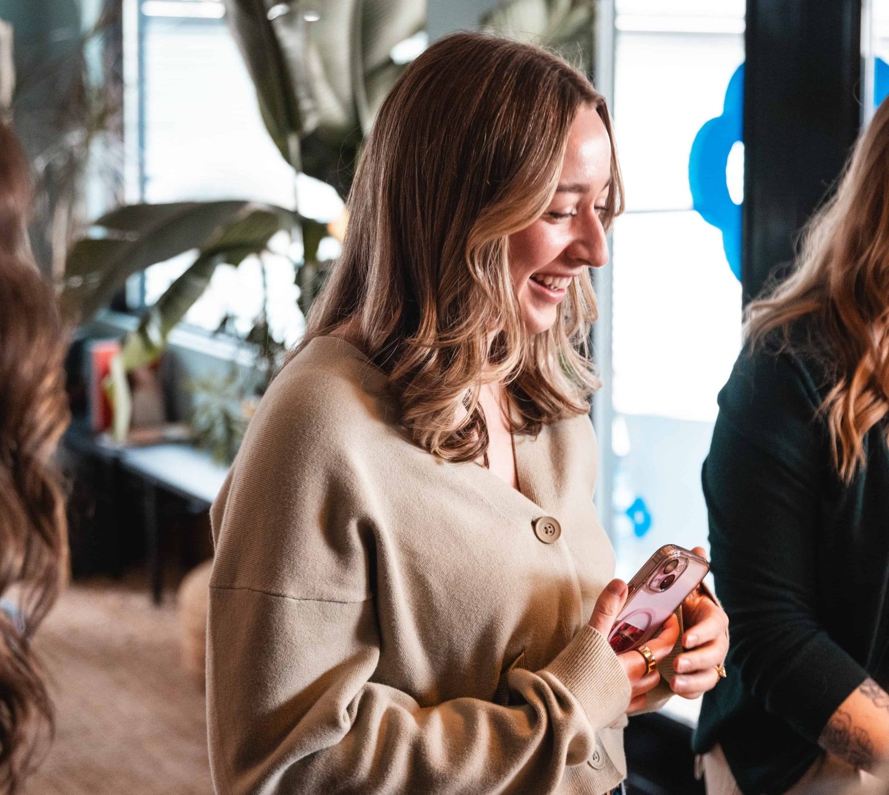 Woman with wavy hair smiles whilst holding a mobile phone indoors, standing next to another person.