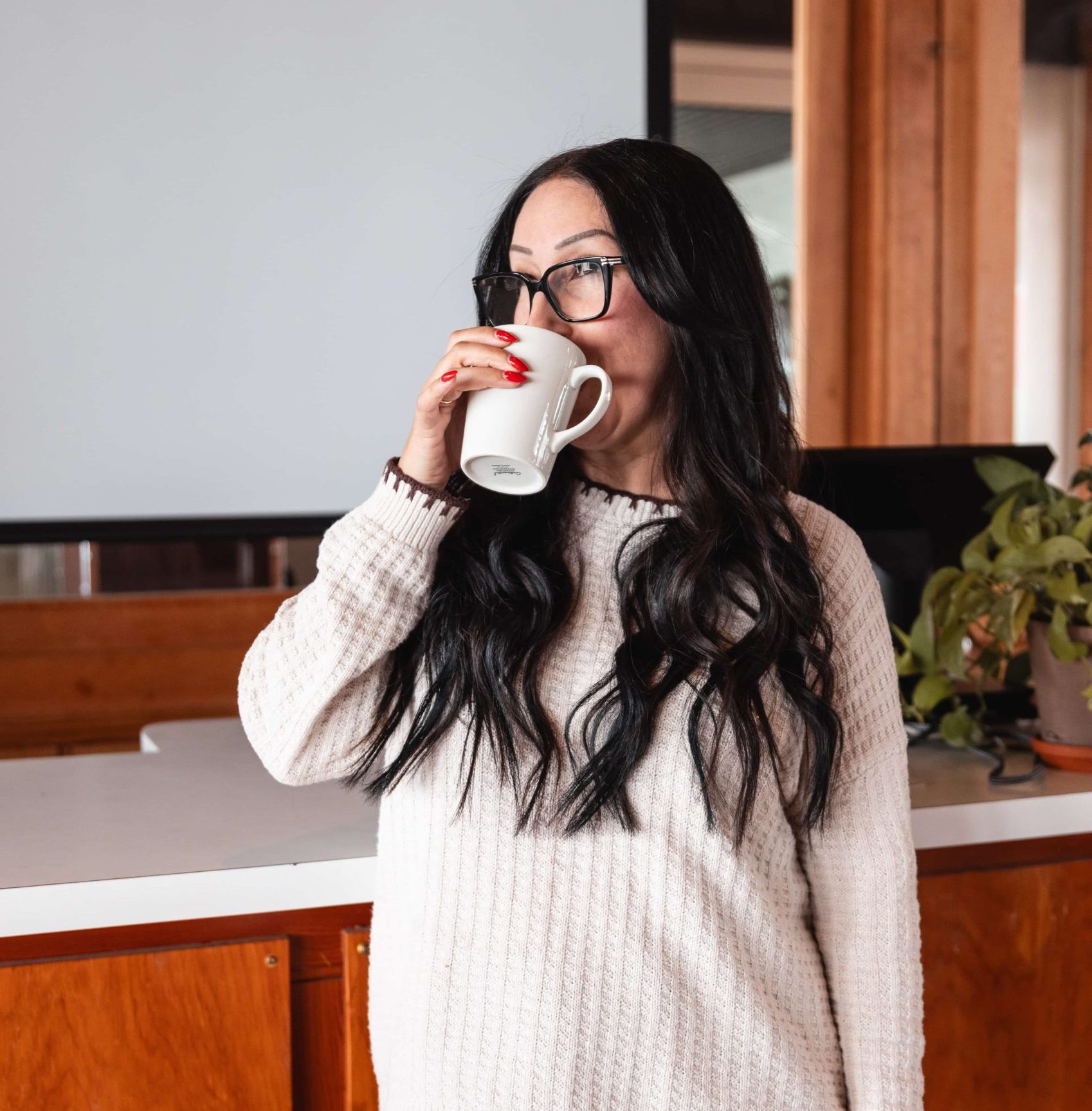 Woman with long dark hair and glasses drinks from a white mug while working remotely for a marketing agency.