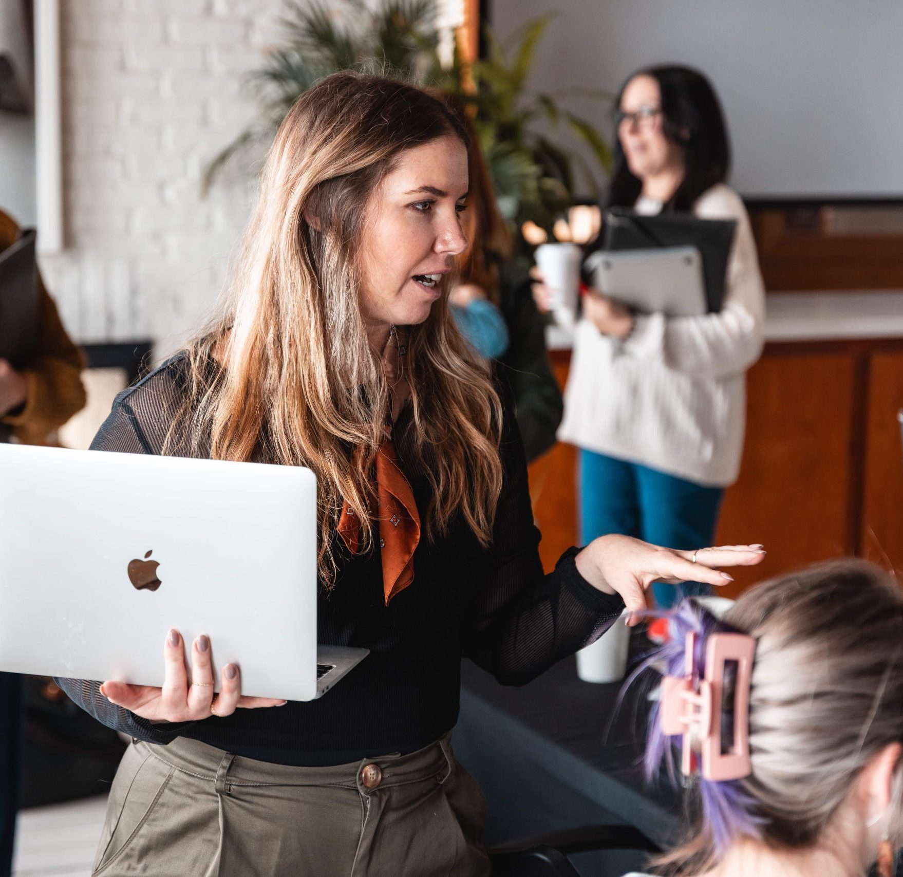 Woman holding a laptop discusses careers with another; two people with devices are in the background.