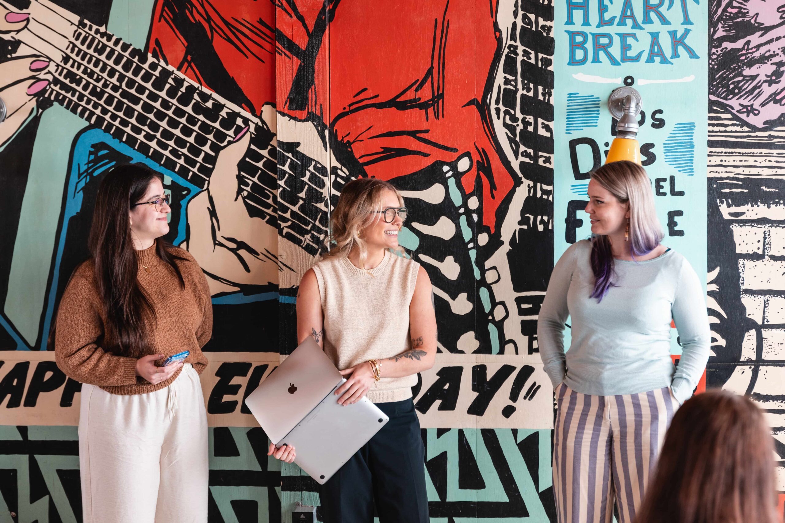 Three women stand talking in front of a colourful mural, one holding two laptops, all appear engaged in conversation.