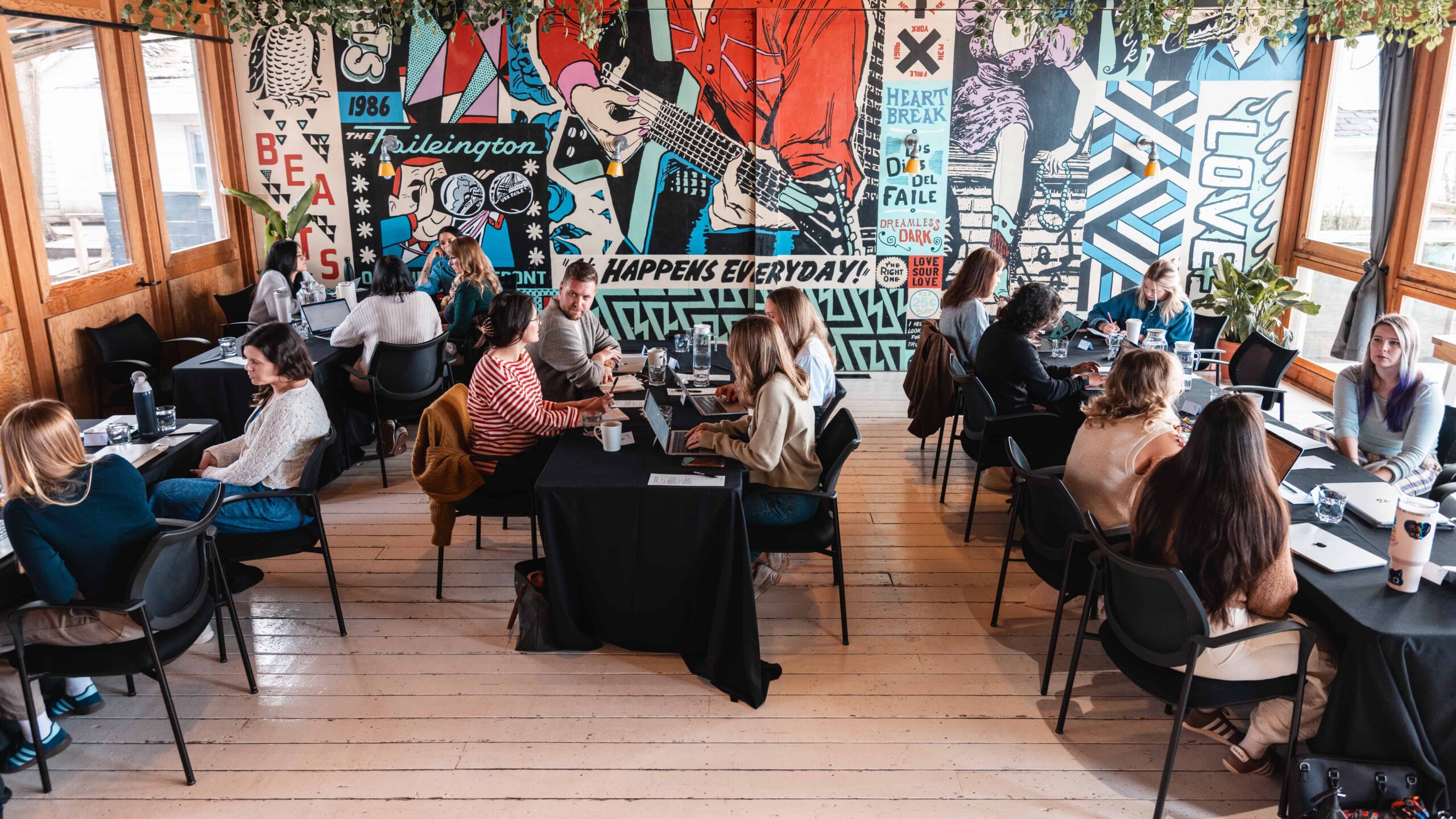 People seated at tables in a bright room, having conversations in front of a colourful mural on the wall.