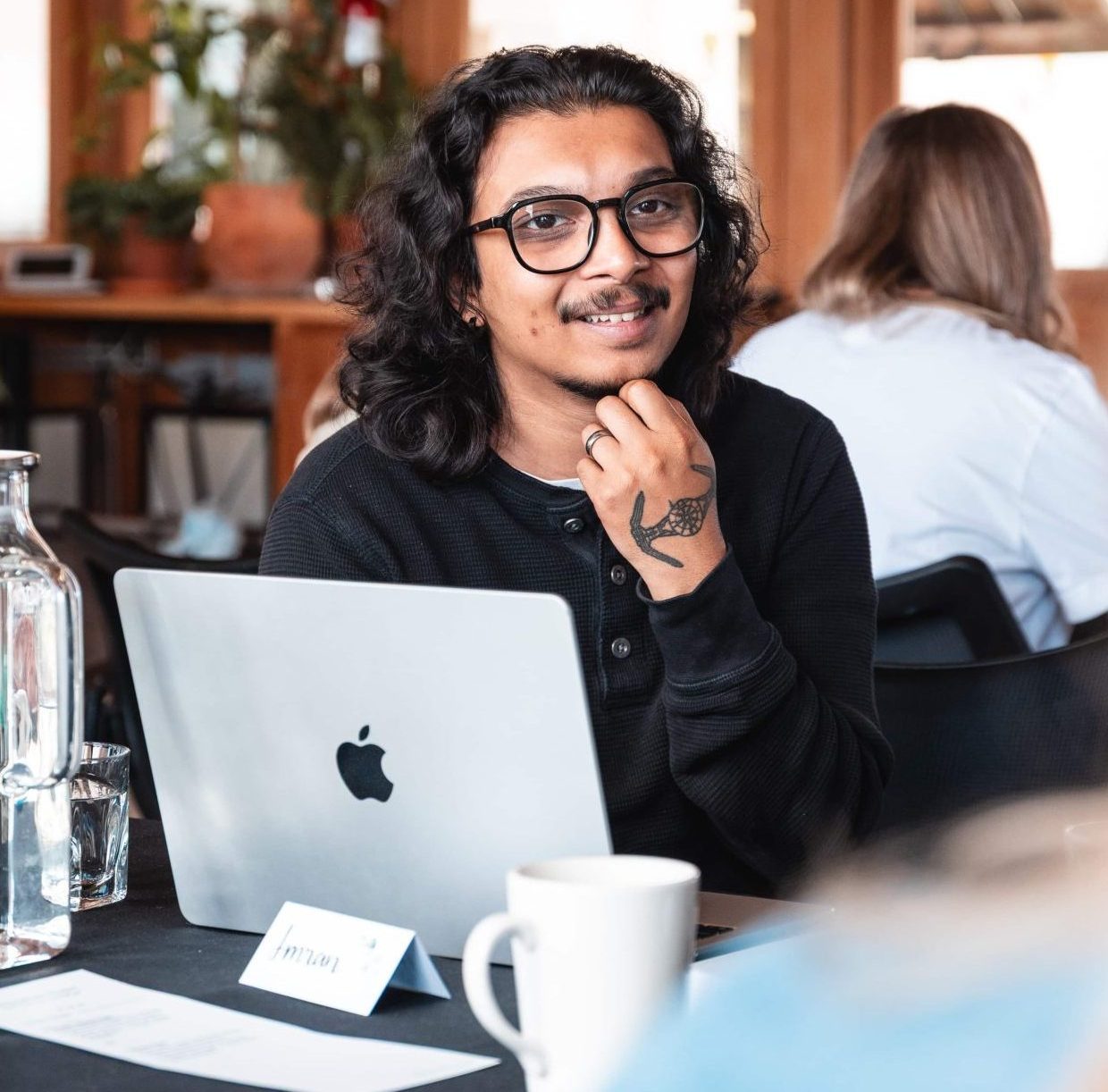 Person with long hair and glasses sits at a table with a laptop, coffee mug, and water bottle, smiling at the camera.