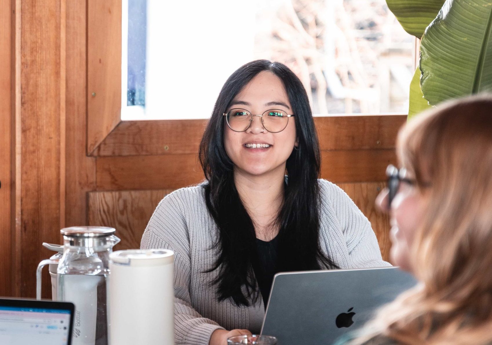 Woman with glasses sits at a table with a laptop, smiling, in a marketing agency’s wooden room with natural light.
