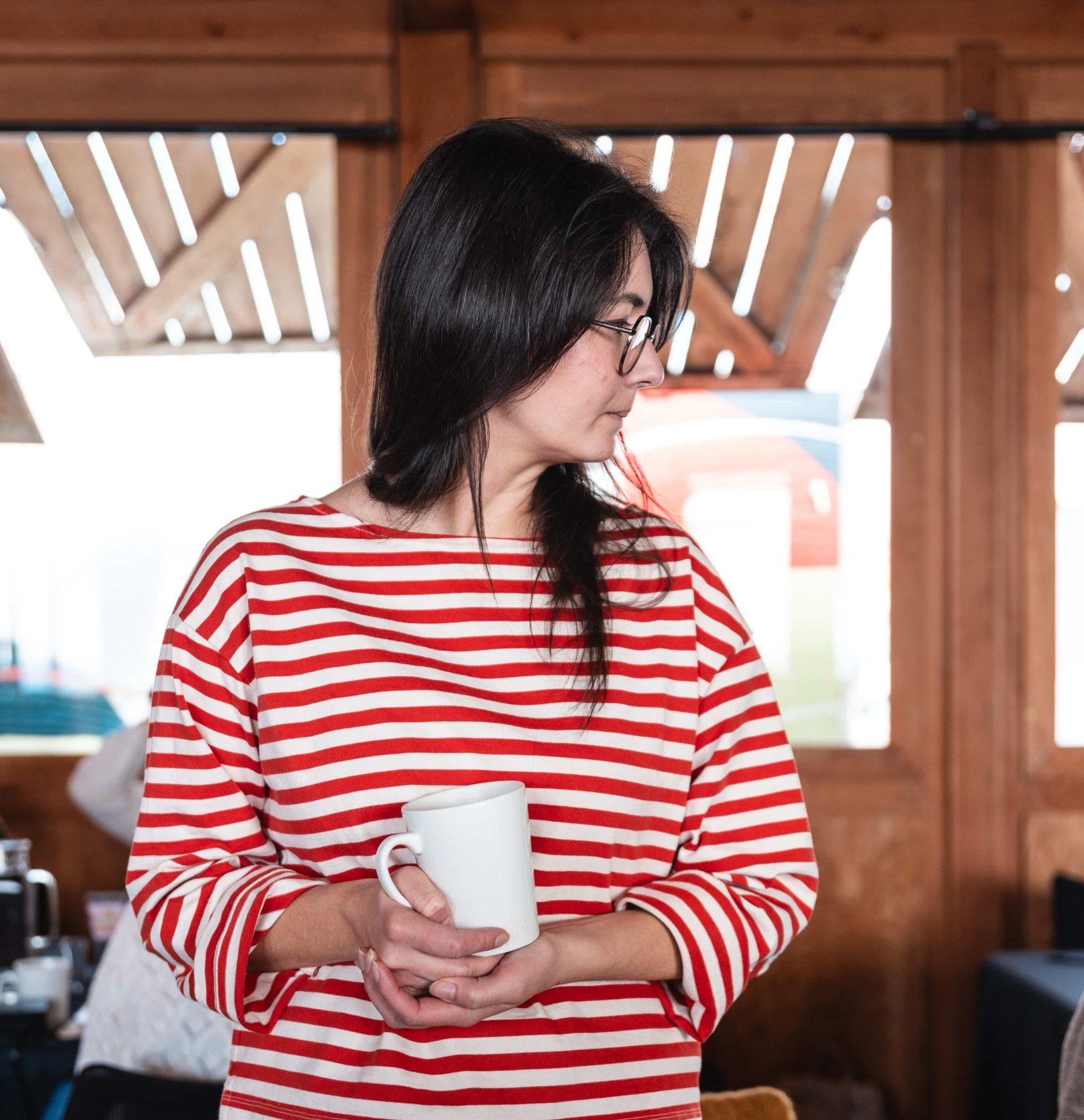 Woman in a red and white striped shirt, mug in hand, ponders ideas for her marketing agency in a wooden interior space.