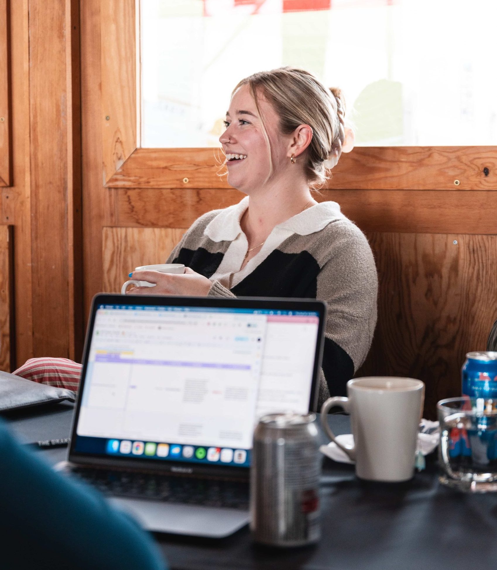 Woman smiling, holding a cup at a table in a marketing agency, surrounded by laptops, mugs, and drinks in bright light.