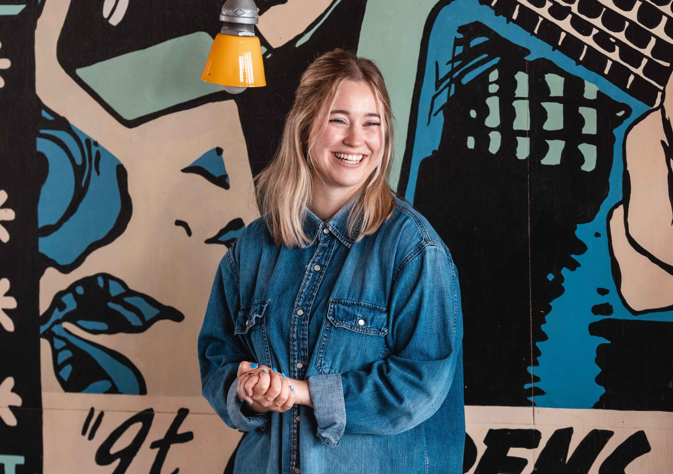 A woman in a denim shirt smiles before a vibrant mural. Have questions? Contact us for more info!.