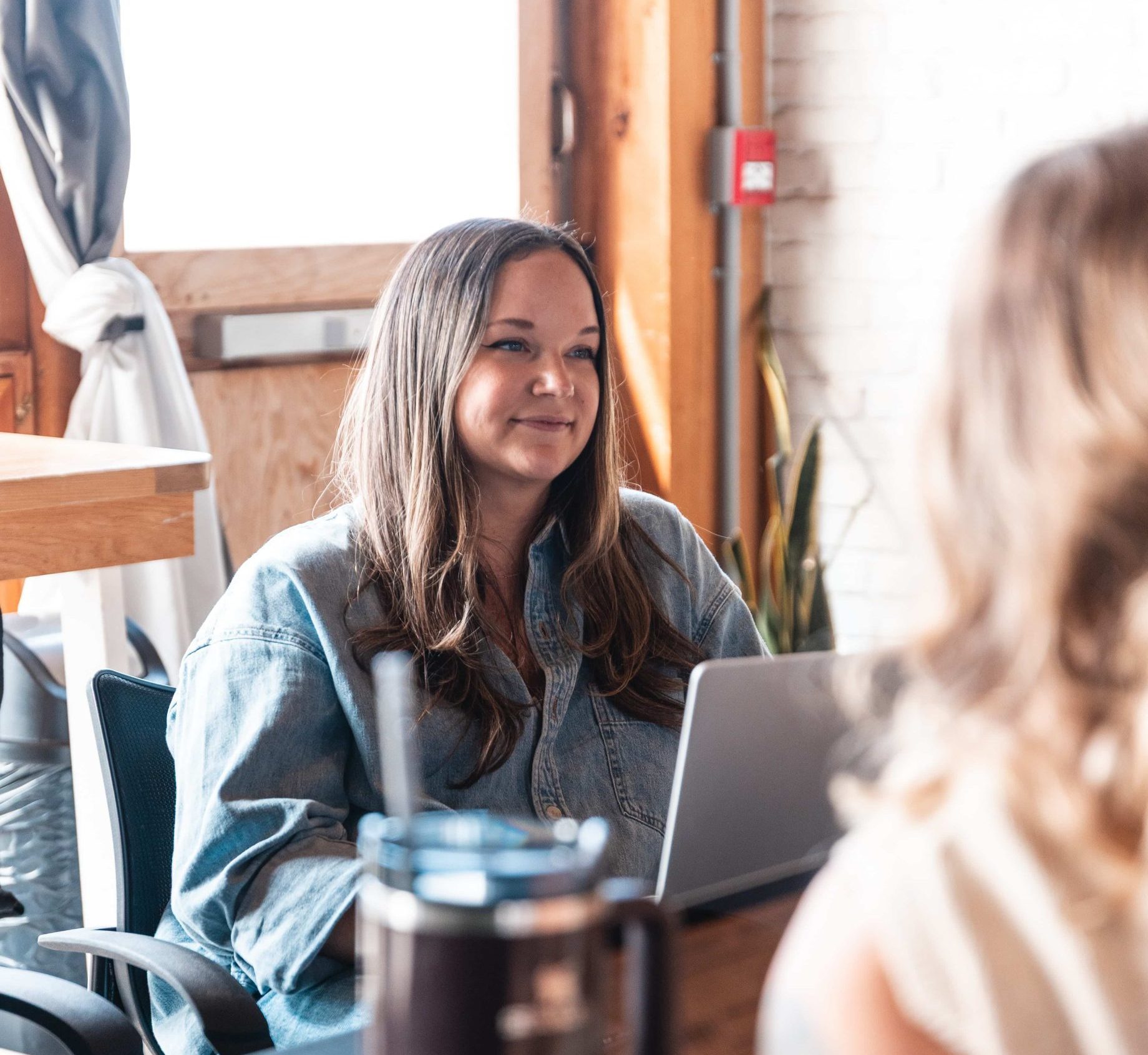Woman with long hair at a desk, smiling as she works for a marketing agency in a bright room with wooden and brick walls.