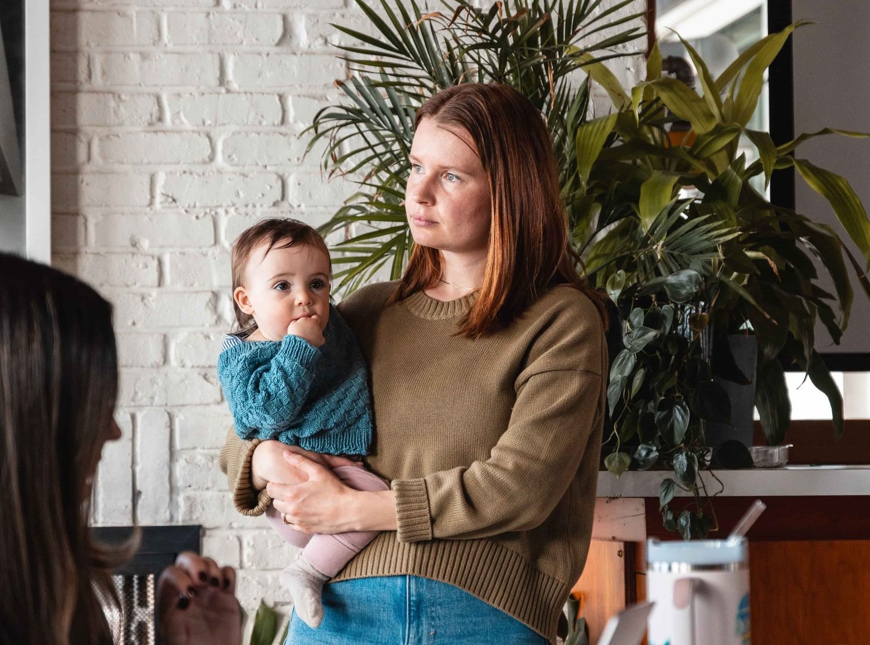 Woman holding a baby stands indoors near plants, looking to the side—perhaps pondering her next marketing agency move.