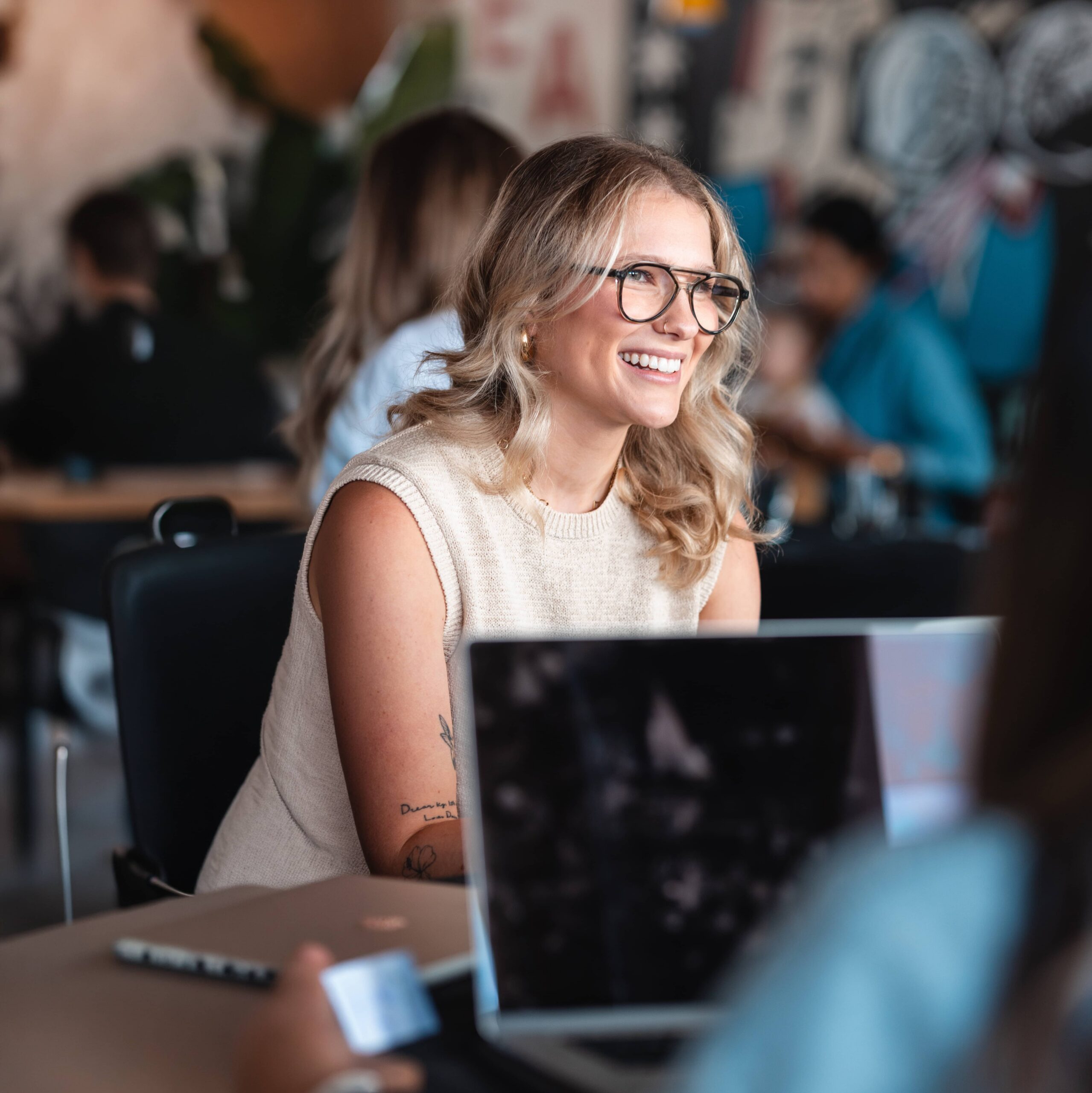 Woman with glasses smiling whilst sitting at a table with a laptop in a casual, busy workplace.