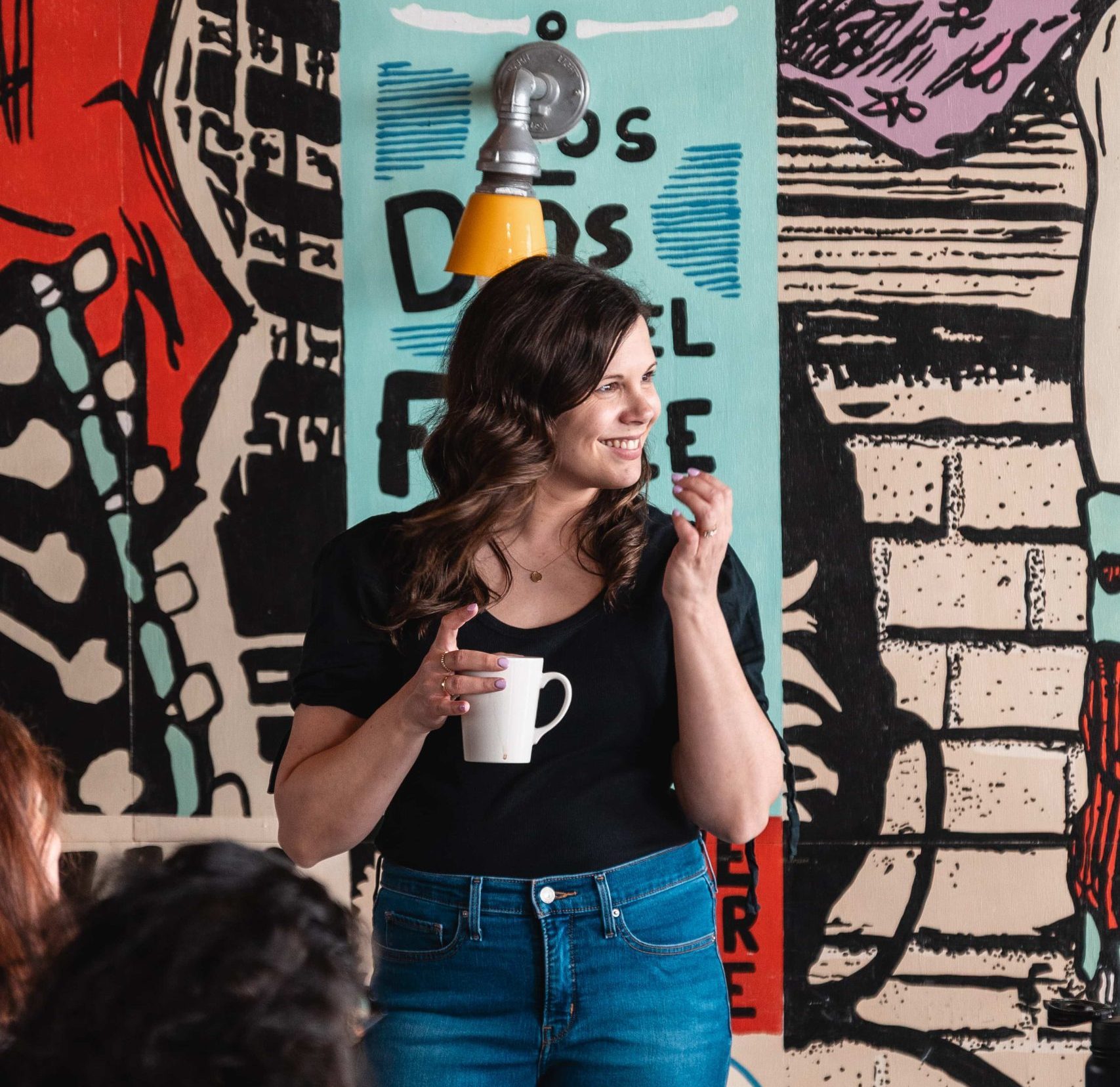 A woman in a black shirt holds a white mug and smiles, standing in front of her marketing agency’s colourful mural.