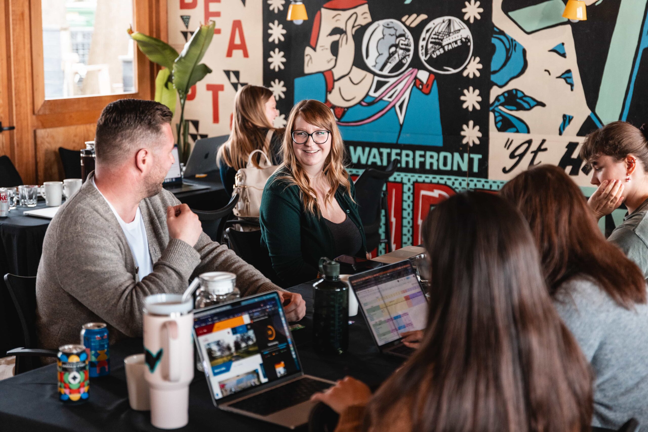 Group of people sit around a table with laptops, talking and working in a colourful, mural-decorated room.