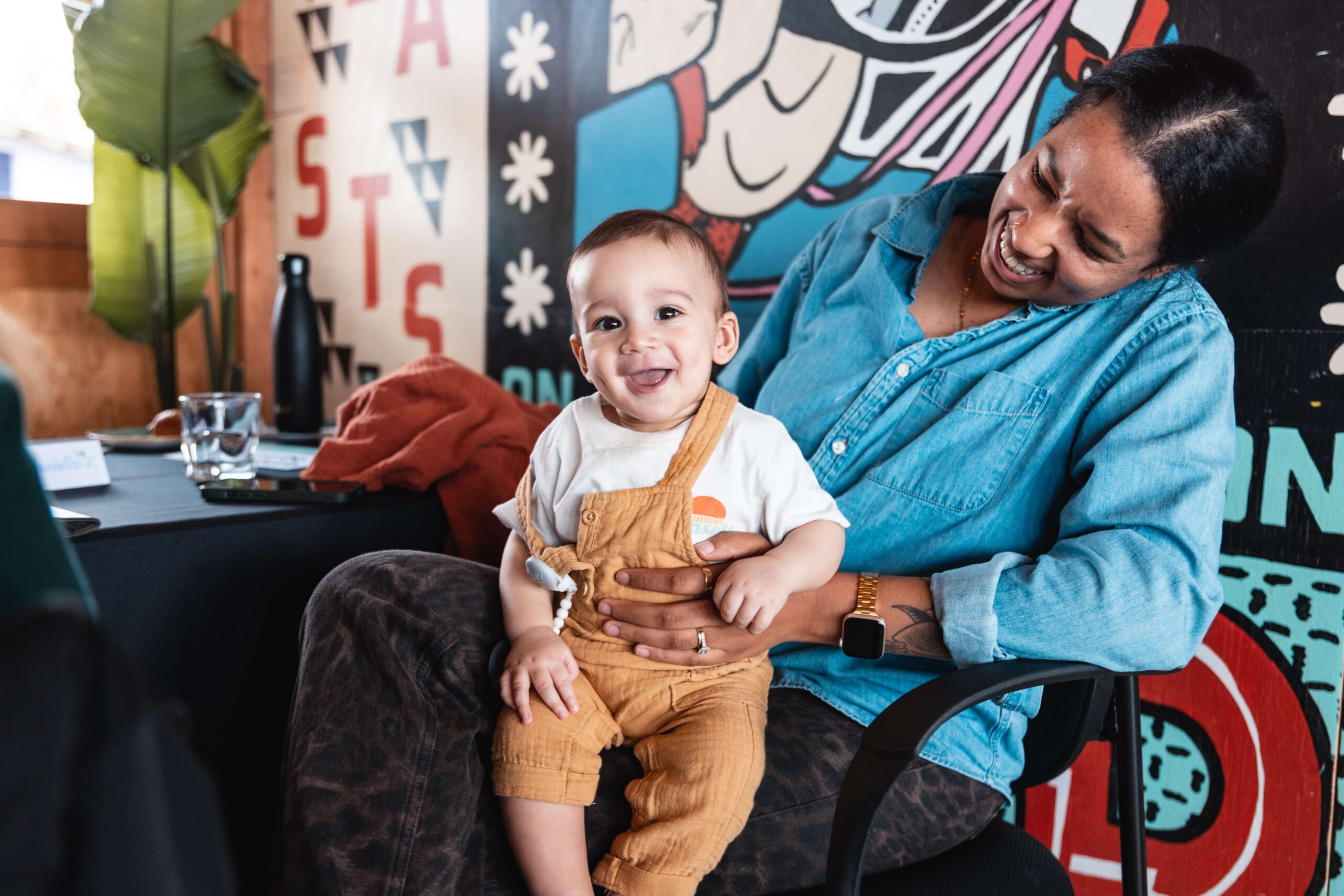 Smiling woman sitting in a chair holds a happy baby on her lap in a colourful, art-filled indoor setting.