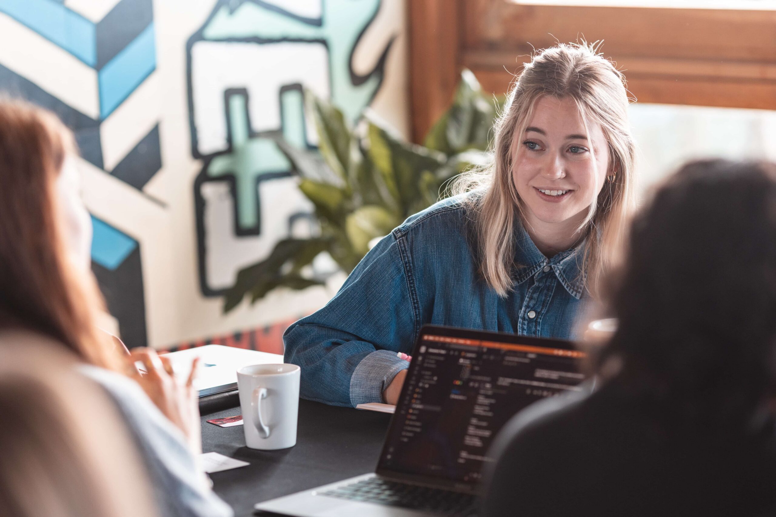 Three people have a discussion at a table with laptops and coffee mugs in a brightly lit, modern workplace.